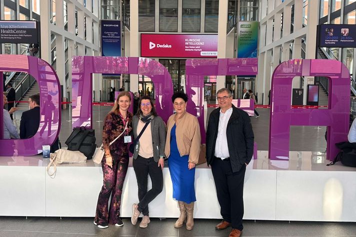 Vier Menschen stehen am Eingang einer Messehalle, im Hintergrund die mannshohen Buchstaben DMEA.