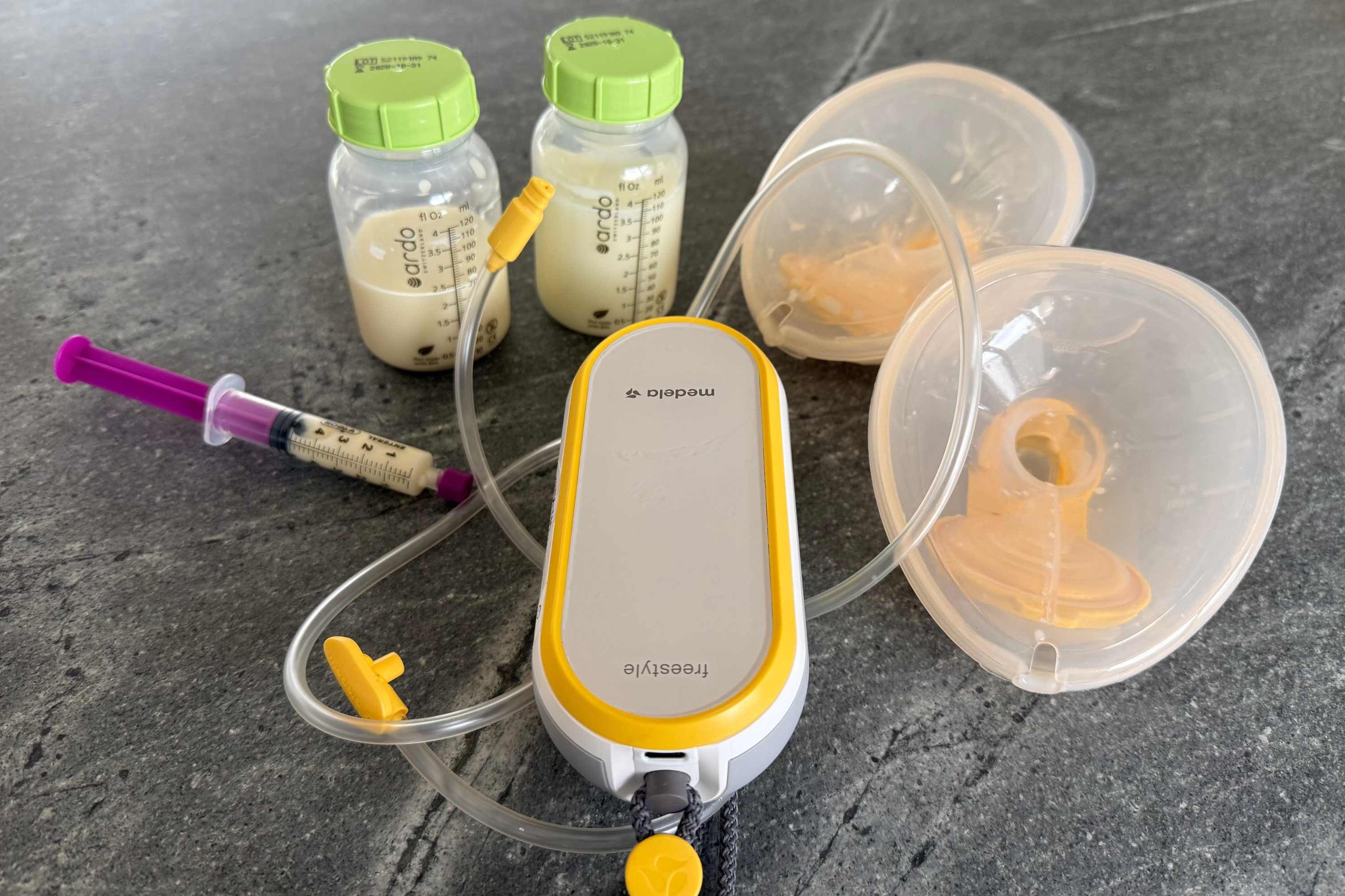 Frauenmilchspende: Auf einer grauen Fläche stehen zwei Fläschchen mit Frauenmilch befüllt, eine Spritze und ein mobiles Milchpumpengerät. Alle diese Dinge benötigt man zur Frauenspende. mit angeschlossenen Brustaufsätzen.