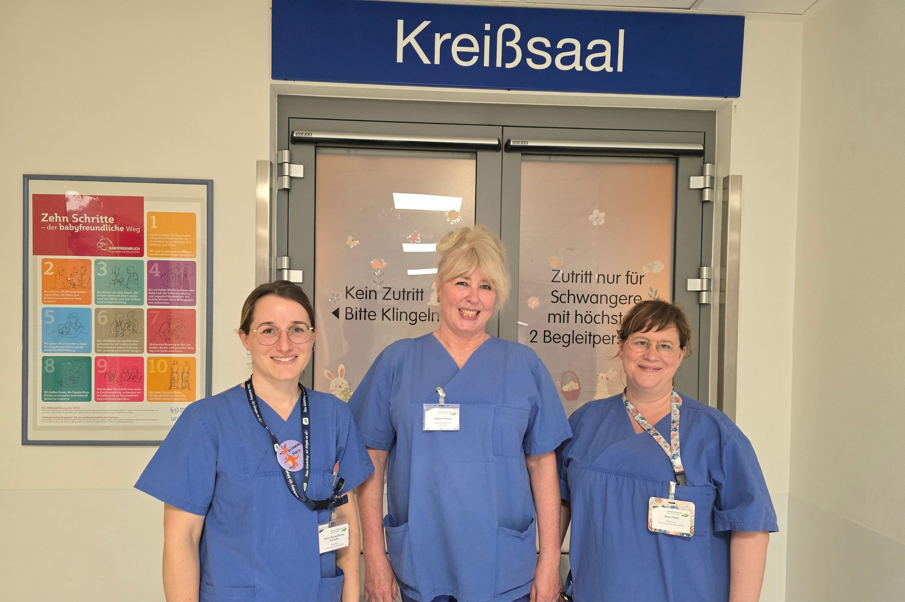Drei Hebammen stehen vor der Tür zum Kreißsaal und lächeln in die Kamera.