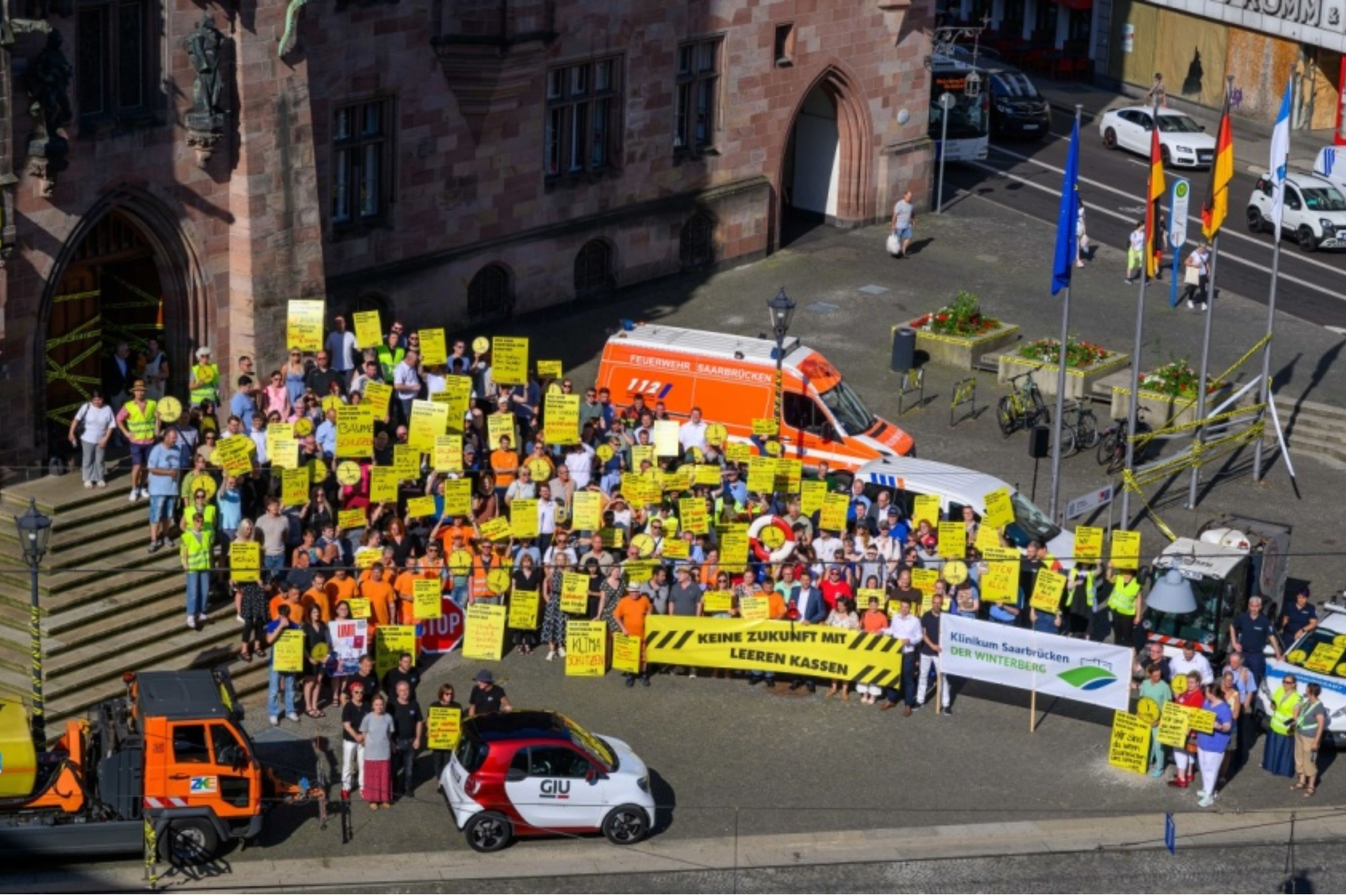 Ein aus der Luft fotografiertes Bild einer Demonstrationsgruppe vor dem Rathaus mit vielen Fahrzeugen (MÜll, Feuerwehr) und vielen gelben Warnschildern.