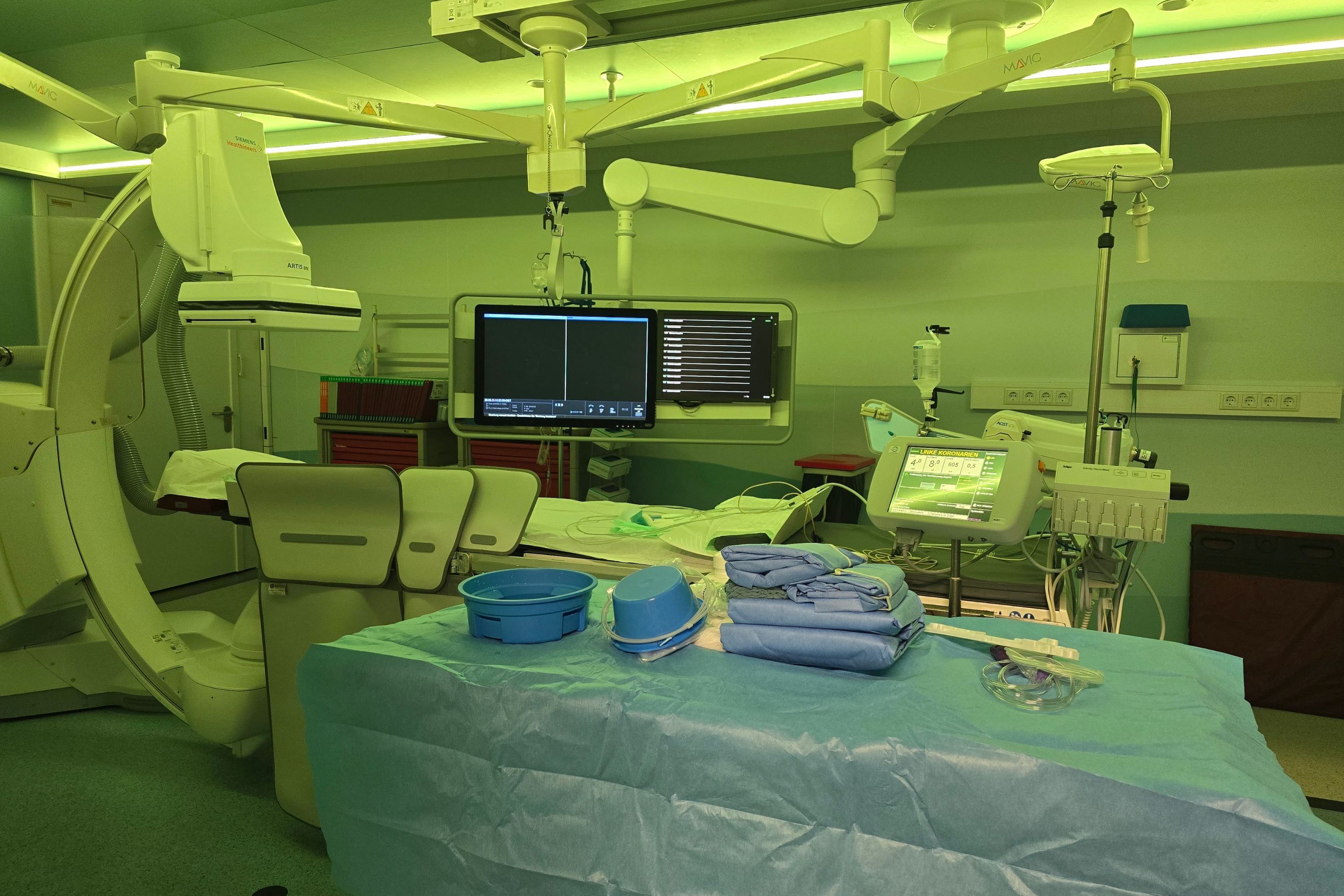 Das Foto zeigt das Herzkatheterlabor von innen, mit grünlichem Licht, aber ohne Patient und ohne Ärzte, nur der Behandlungstisch und die Monitore und die vorbereiteten sterilen Instrumente.