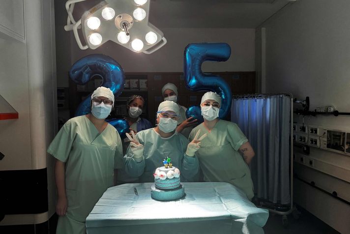 Fünf Operationstechnische Assistenten und Assistentinnen im Operationssaal mit einer grünen Torte und zwei Luftballons in Form von Zahlen, einer zwei und einer fünf.