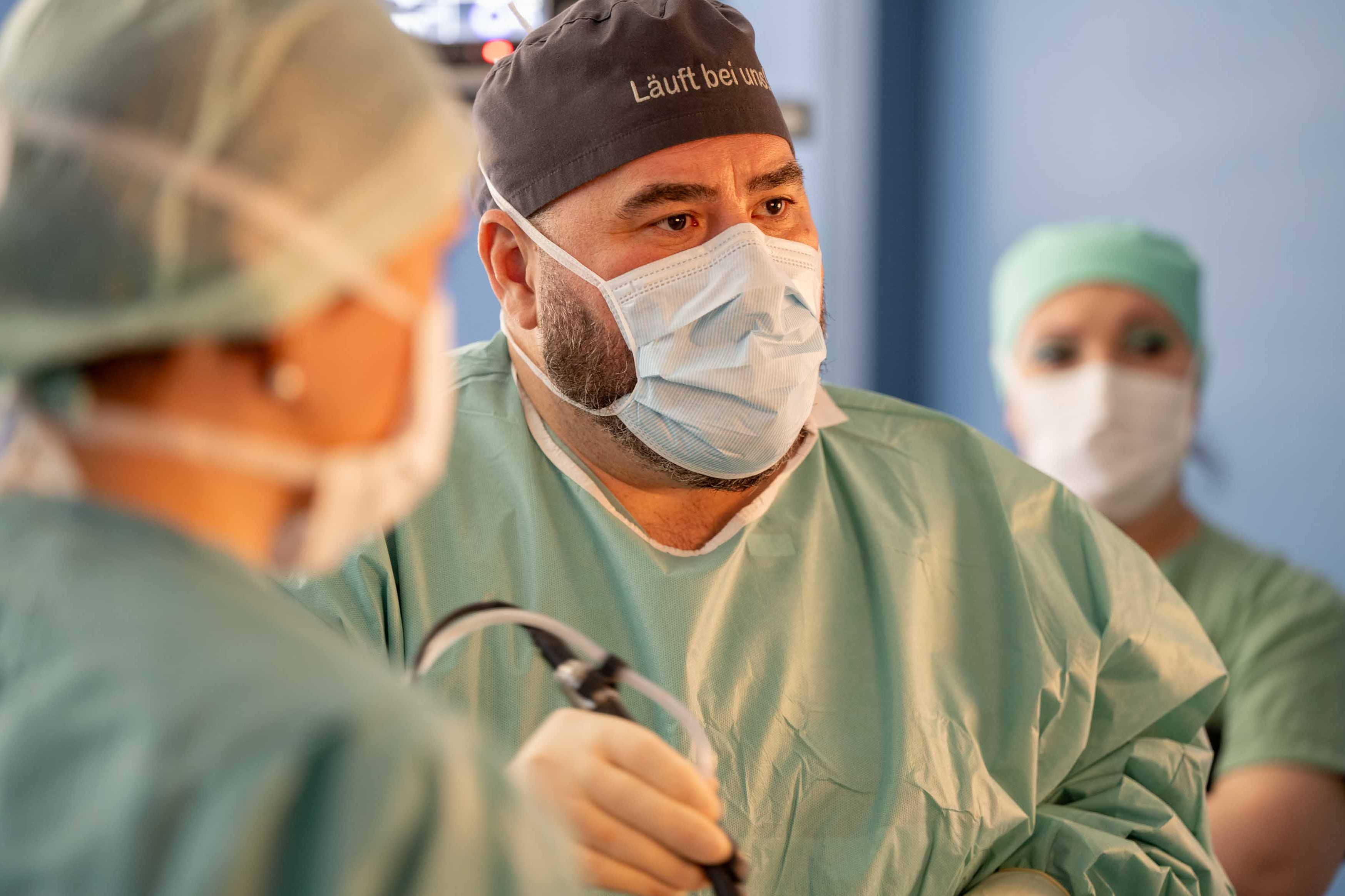 Dr. Dr. Habil Gregor A. Stavrou während einer Operation, mit steriler OP-Kleidung, Mund-Nasen-Schutz und OP-Haube mit Aufschrift "Läuft bei uns."
