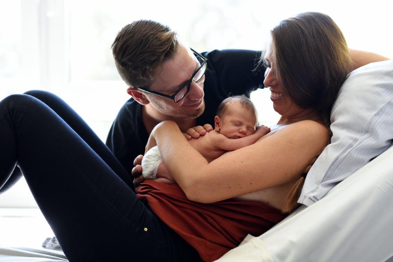 Familie mit Baby, Mutter hat Neugeborenes auf der Brust, Vater schaut neben dem Bett auf das Baby