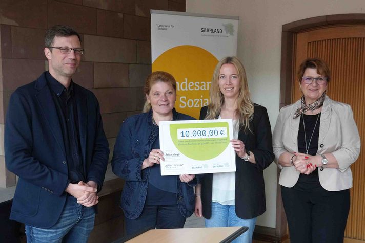 Die zwei Beauftragten für Betriebliches Eingliederungsmanagement nehmen einen symbolischen Check entgegen im Landesamt für Soziales vom Projektbeauftragten und von Bettina Altesleben.