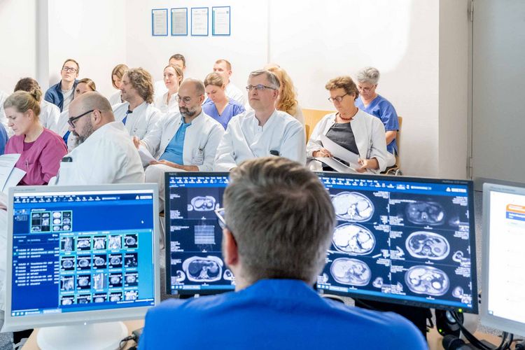 Medizinisches Fachpersonal nimmt an einer Tumorkonferenz im Klinikum Saarbrücken teil. Im Vordergrund steht ein Präsentator vor drei Computerbildschirmen, auf denen medizinische Daten und Bildmaterial angezeigt werden. Die Teilnehmenden tragen Arbeitskleidung und befinden sich in einem modernen Konferenzraum mit klinischem Ambiente.