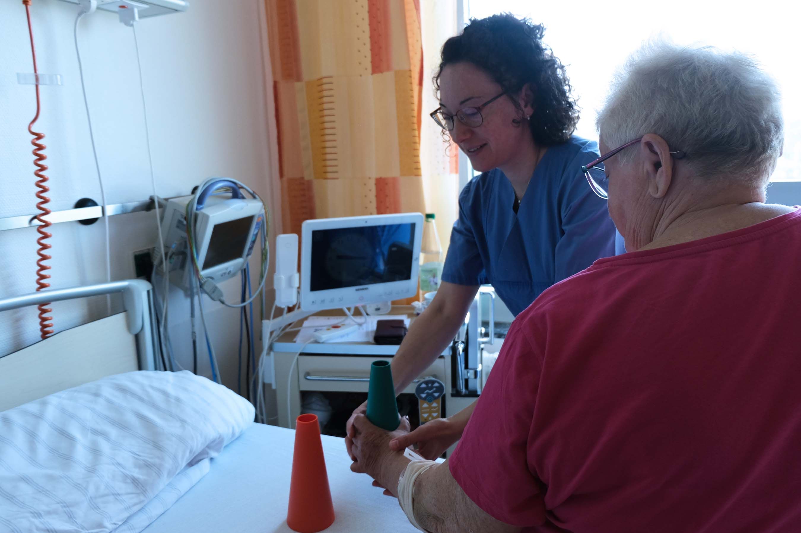 Ergotherapeutin Angela Golesne übt mit einer Patientin nach einem Schlaganfall am Bett Alltagsbewegungen, wie das Bewegen von kleinen Kegeln von der linken zur rechten Körperseite