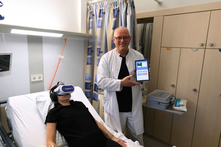 Auf dem Foto sieht man eine Patientin, die eine VR-Brille trägt. Nebendran steht Psychoonkologe Wolfgang Merda und hält ein Tablet in der Hand